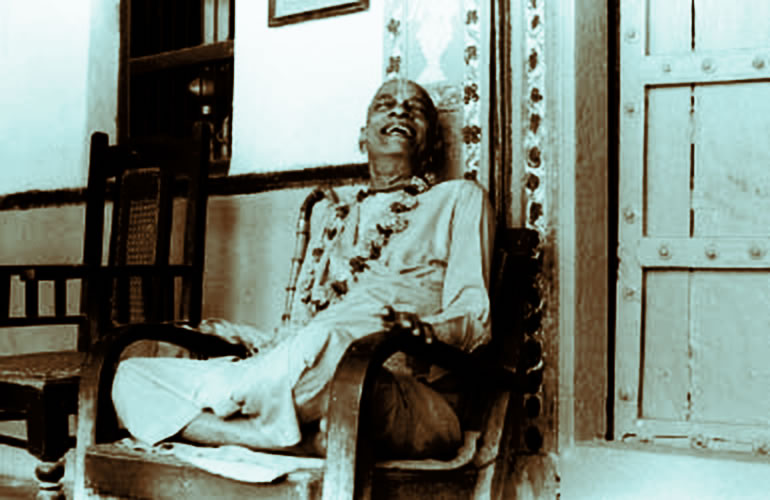 Prabhupada sitting in an old chair in India laughing.